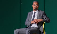 Vaughan Gething, dressed in grey suit with open jacket over white shirt, sitting with hands together on a bright yellow chair against a dark green background