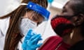 A younger Black nurse with long thin braids, wearing a face shield, face mask and rubber gloves gives a shot in the elbow to an older Black woman wearing a red and black facemask and red top.