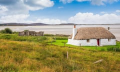 A traditionally built white cottage with thatched roof, next to a turquoise bay, with cloudy skies above.