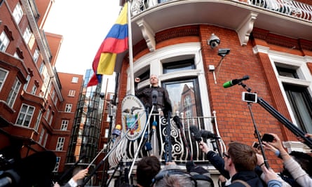 Assange stands on a balcony with the Ecuador flag flying from it and people standing below holding up microphones and cameras on poles