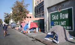 Tents line a sidewalk alongside a building with a green and white painted sign that says 'Skid Row, City Limit'.
