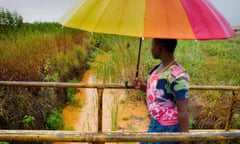 A young woman carrying an umbrella walks along a bridge over a muddy orange-brown stream