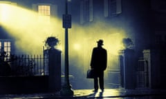 still from the exorcist of a man standing outside a house with yellow light spilling out into the night