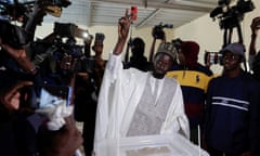 A man wearing a white robe over a grey suit gives a peace sign while standing in front of a ballot box