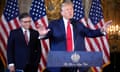 Two men in blue suits and red ties stand at a podium in front of a row of American flags