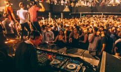 A crowd of dance music fans on the dancefloor at Amsterdam club De Marktkantine. The image is taken from behind the DJ booth, where a DJ is using digital decks to play music.