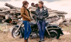 Jodie Comer and Austin Butler standing beside a bike in front of a pile of logs in Bikeriders