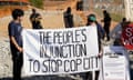 Two young people in red dusty area between chain-link fences, with police officers in the background, hold white cloth banner between them that reads The People's Injunction to Stop Cop City.