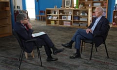 Two white men sit in adjacent chairs with legs crossed, talking.