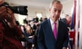 Nigel Farage in pink tie looking downcast with photographer close to his right and seated audience in background