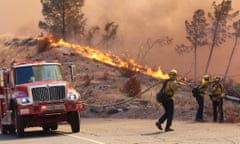 firefighters stand on a road with a red truck while a fire blazes in the background along a line of trees