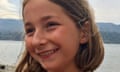 close-up photo of Martha smiling; she has collar-length fair hair tucked behind her ears and is standing in front of a lake or stretch of water with hills seen in the background; her hair is blowing a little in the breeze.