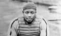 A black-and-white photo of a young, Black baseball catcher, crouching and looking at the camera as he wears a backwards baseball cap, a protective vest, and a catcher's mitt on his left hand, resting his forearms on his thighs.