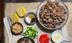 A dish of ful medames (Egyptian stewed fava beans) surrounded by bowls containing chopped cucumber, chopped tomatoes, crushed garlic, sauteed onions, plus lemon halves.