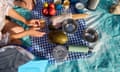 Aerial shot of a picnic with colorful fruits, bowls, and a thermos on a checkered blanket. Hands are seen arranging food, ready for outdoor dining.<