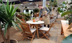 A waiter adjusts the setting of a small round table, in a light room filled with plants