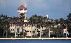 Large salmon-pink structure with red roof tiles and a tower seen among dozens of palm trees from across a blue bay.