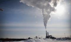 A bright yet hazy sky, with the sun peeking around a plume of white smoke rising from one of two tall smoke stacks, with low snowy fields in the foreground.