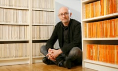 Nicholas Royle sitting crosslegged between shelves of colour-coded paperbacks.