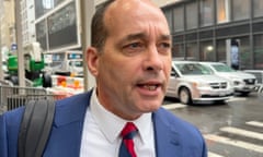 Close-up outdoor shot of a man in a city intersection. He is white, middle-aged, mostly bald, wears a blue suit and a striped red and blue tie, has a bag slung over one shoulder, and appears to be speaking.