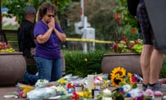 A woman kneels, her hands held together in prayer, in front of a cluster of flower bouquets.