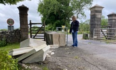 A person stands by a stone plinth with a crack in it next to some other pieces of stone beside a cattle grid and gateway