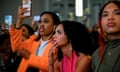 Three young Black women, one is bright orange, one in bright pink, and one in olive, watch a live video on a cellphone.