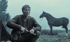 Mads Mikkelsen in ragged clothes and holding a pistol, looking serious, sat on his haunches outside a tent in the mist, a horse in profile in the distance