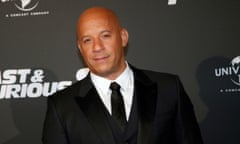 A mixed-race man with a totally bald head, very muscular and stocky, sort of smiles wearing a black suit and tie, and white shirt, in front of background that has film logo on it, in black and white.