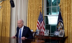 man wearing suit and blue tie sits at table in front of yellow curtains, two flags and photo frames