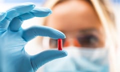 Young attractive female scientist holding a red transparent pill<br>Young attractive female scientist with protective eyeglasses and mask holding a red transparent pill with fingers in gloves in the pharmaceutical research laboratory