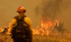 person wearing yellow shirt and red hat and carrying black backpack looks at flames in front