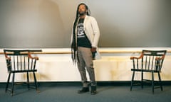 Black man with very long dreads, wearing black T-shirt, cream blazer, and gray khakis poses in front of long chalkboard between two chairs.