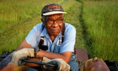 A man wearing a hat, denim shirt, and gloves sits on a tractor on farmland