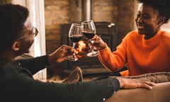 Joyful young couple cheering with glasses of red wine while celebrating their anniversary<br>Candid shot of joyful young couple relaxing on the sofa, in front of a cozy fireplace, smiling at each other and cheering with glasses of red wine while celebrating their anniversary.