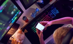 Top-down view of a person holding their phone over a lit-up keyboard with two screens in front of them and junk food on the desk