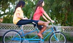 Two women riding a tandem bike while the woman on back surfs the internet on a laptop