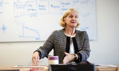 Amanda Spielman photographed behind a desk with a whiteboard behind her, in what looks like a classroom; she has chin-length blond hair and is wearing a collarless grey tweed jacket, and is gesturing with her hands