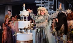 Samuel Boden, arms raised, dressed as a beaming bride, with a three-tier wedding cake close by, in the title role of Platée at Garsington.