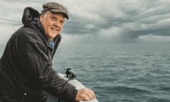 Bryn Terfel in waterproof and flat cap on a boat at sea, grey storm clouds in the distance