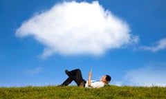a man lying on grass under blue skies and a big fluffy cloud, looking delightedly at his tablet