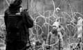 A black and white image of a child behind razor wire looking up at armed border guard.