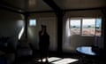 A woman stands in a building with a table, chair, wood beams and two windows.