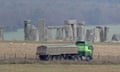 A lorry in the foreground passes the freestanding stone structures of Stonehenge, which is in a field behind it