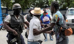 An armed police officer has his hand on a man's arm who is being led away by another man. Bystanders protest in the background.