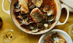 Hake with clams, chorizo and manzanilla in an iron pot with handles on a tablecloth next to a bowlful of it, a plate of torn bread, a bowl of clam shells and a wine glass