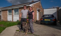 Graham and Lisa stand with their arms round each other with their dog on the driveway of. a brick bungalow