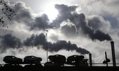 Smoke rises from a factory as a truck loaded with cars crosses a bridge