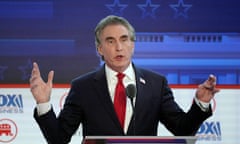 A man wearing a suit, white shirt and red tie gestures as he speaks in to a mic.