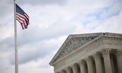 Taken from below, the top of the white-pillared US supreme court building, with a cloudy sky beyond it and a US flag at the top of a flagpole.